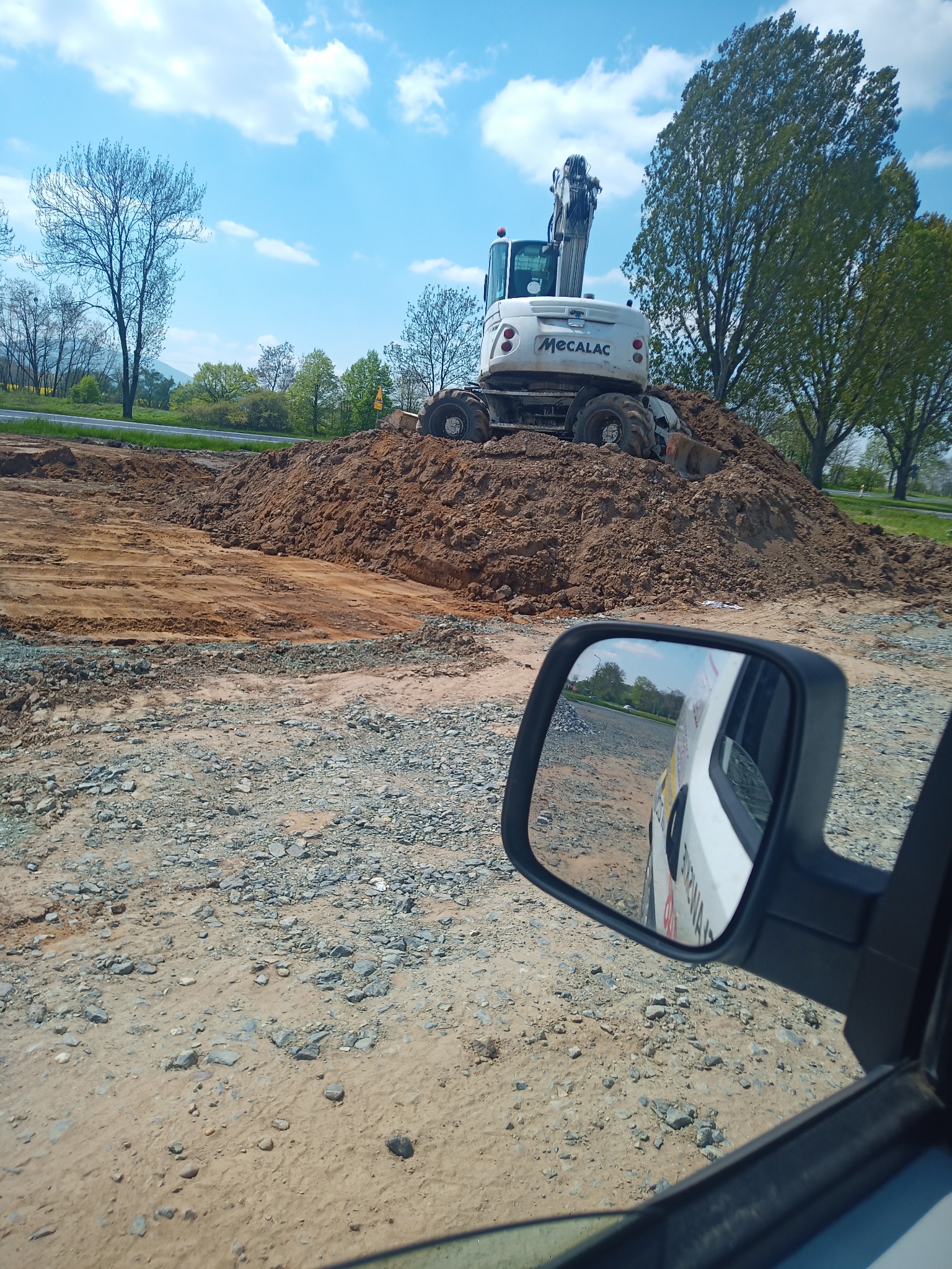 Biała koparka Mecalac na hałdzie ziemi, widok z perspektywy kierowcy z lusterkiem bocznym w kadrze, teren budowy w tle, błękitne niebo z chmurami.