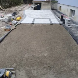 Przygotowany teren pod ścieżkę z kostki brukowej, częściowo ułożona kostka, widoczny ubijak wibracyjny i zapas kostki obok budynku.