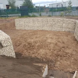 Gabionowe ściany oporowe tworzące taras na nierównym terenie, widoczne metalowe kosze wypełnione kamieniami, ziemia przygotowana pod dalsze prace.