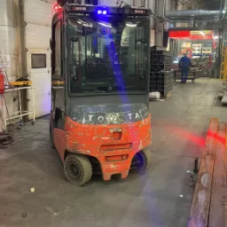 Używany wózek widłowy Toyota z włączonymi niebieskimi światłami ostrzegawczymi, widoczne ślady użytkowania na pomarańczowej obudowie, w tle hala produkcyjna z elementami metalowymi i pracownikiem.