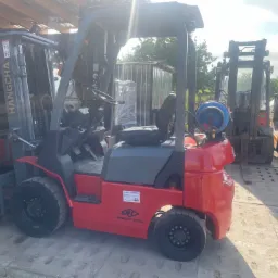 Czerwony wózek widłowy marki Hangcha z butlą gazową, stojący na betonowej nawierzchni w zadaszonym magazynie, z innymi wózkami w tle.