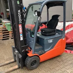 Nowy wózek widłowy Toyota Trigo 18, szaro-pomarańczowy, z kabiną i czarnym fotelem, stojący na kostce brukowej przed budynkiem.