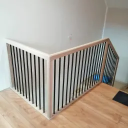 Fragment jasnej drewnianej balustrady schodowej z czarnymi, pionowymi szczeblami, zamontowanej przy białej ścianie i drewnianej podłodze.