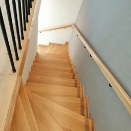 Widok z góry na jasne, dębowe schody z czarną metalową balustradą po lewej i drewnianą poręczą po prawej stronie, przy szarej ścianie.