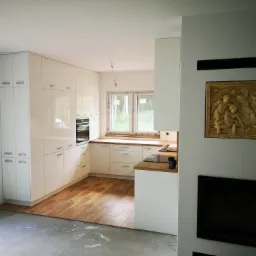 Nowoczesna biała kuchnia z drewnianym blatem i podłogą w trakcie wykańczania, widoczny fragment surowej podłogi, dekoracyjny relief nad kominkiem.