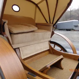 Wnętrze eleganckiej, drewnianej bryczki z beżową tapicerką i baldachimem, widok z boku na siedzenie i koła, na zewnątrz widoczny krajobraz z drzewami i przyczepą.