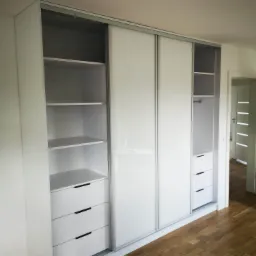 Biała szafa wnękowa z przesuwnymi drzwiami i szufladami, widoczne p&oacute;łki i drążek na ubrania, parkiet na podłodze, drzwi w tle.