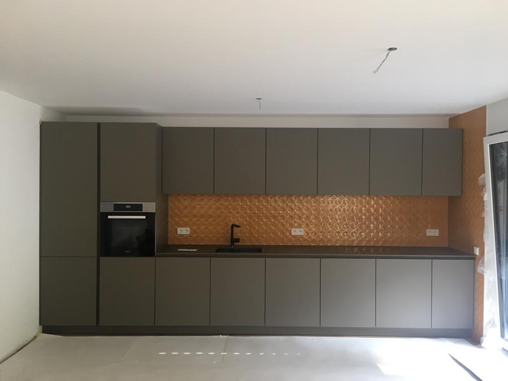 Nowoczesna kuchnia z szarymi szafkami, pomarańczowym panelem dekoracyjnym nad blatem i czarną armaturą, jeszcze w trakcie wykańczania, widoczne kable wystające z sufitu.