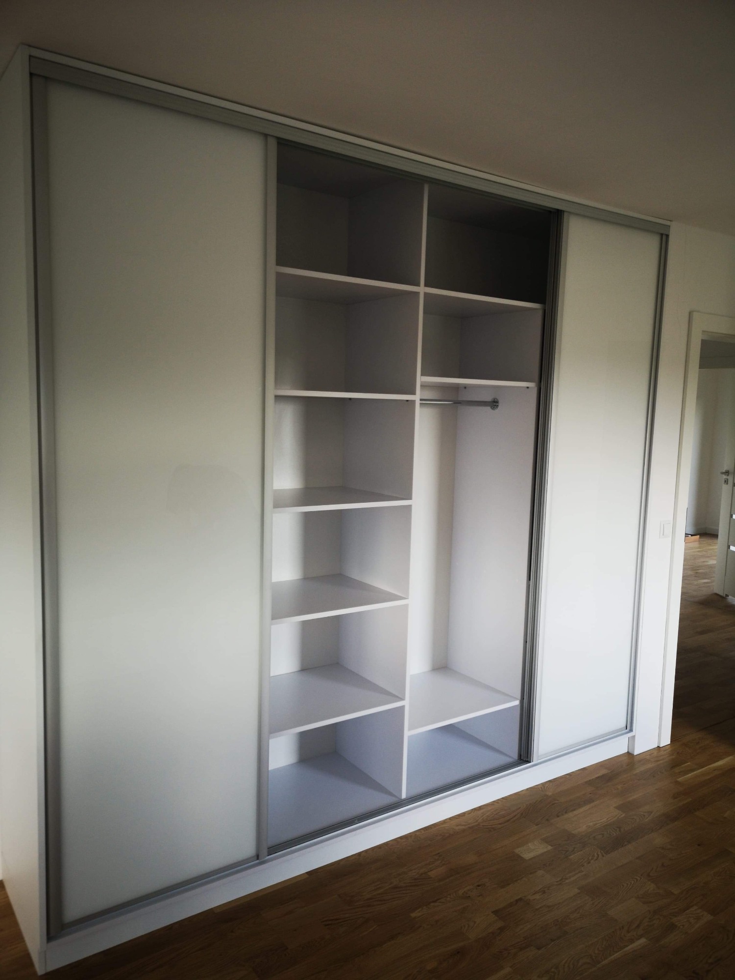 Biała szafa wnękowa z przesuwnymi drzwiami i półkami, widok z przodu na tle drewnianej podłogi.