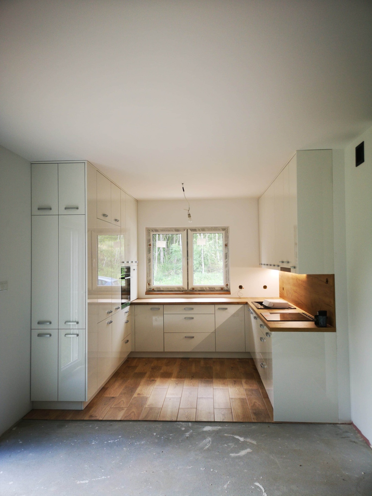 Nowoczesna, biała zabudowa kuchenna z drewnianym blatem i okładziną ścienną, widok na okno i częściowo wykończoną podłogę, surowy stan ścian.