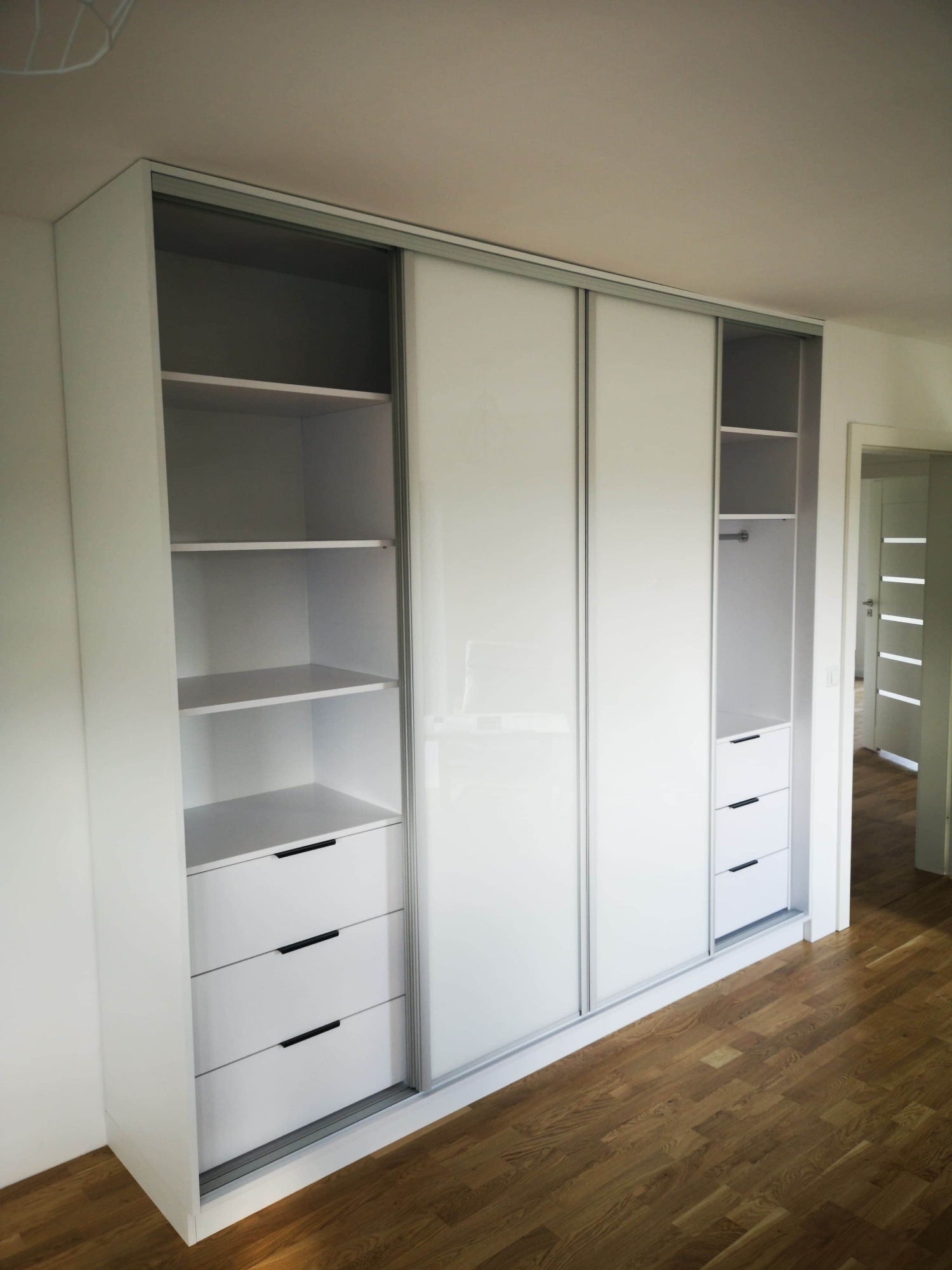 Biała szafa wnękowa z przesuwnymi drzwiami i szufladami, widoczne półki i drążek na ubrania, parkiet na podłodze, drzwi w tle.
