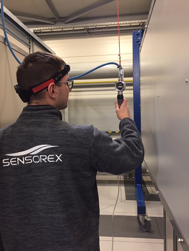 Mężczyzna w okularach i z lampą na głowie, w bluzie z logo Sensorex, dokonuje pomiaru urządzenia zawieszonego na lince w pomieszczeniu technicznym z metalową konstrukcją.