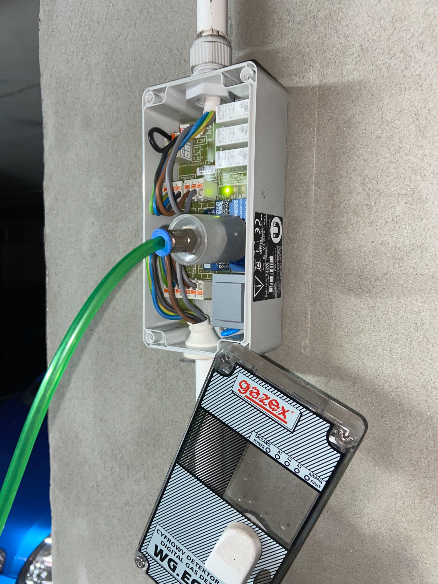 Instalacja czujnika gazu Gazex WG.EG w obudowie, z widoczną elektroniką, przewodami i zielonym wężem doprowadzającym gaz, zamontowana na szarej ścianie.