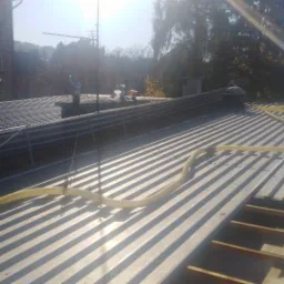 Metalowy dach z widoczną żółtą rurą wentylacyjną i fragmentem odsłoniętej konstrukcji drewnianej, w tle budynek z anteną satelitarną i drzewa.