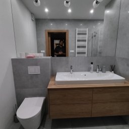 Redesign Bogumił Wilamowski - Nowoczesna łazienka z szarą płytką, drewnianą szafką pod umywalką i wiszącą toaletą; w lustrze odbija się osoba i grzejnik drabinkowy.