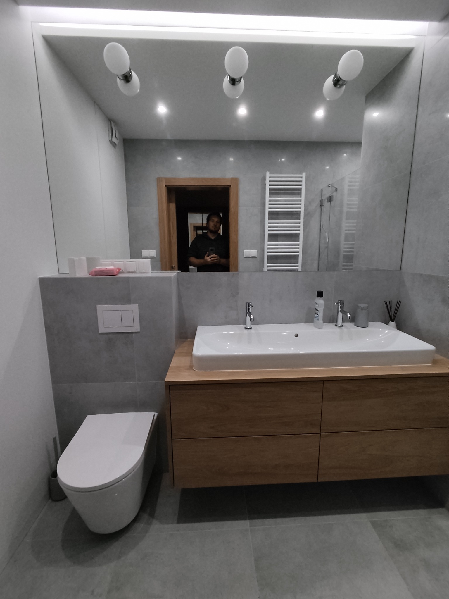 Nowoczesna łazienka z szarą płytką, drewnianą szafką pod umywalką i wiszącą toaletą; w lustrze odbija się osoba i grzejnik drabinkowy.