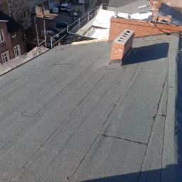 Widok z góry na płaski dach pokryty papą termozgrzewalną z widocznym kominem z cegły i fragmentami innych budynków w tle.