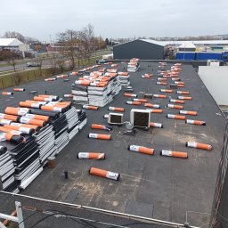 Karol Sitkowski Usługi Budowlane Montażowe Dekarskie - Widok z góry na płaski dach z rozłożonymi rolkami papy termozgrzewalnej marki Bauder w pomarańczowych opakowaniach oraz ułożonymi w stosy płytami izolacyjnymi z polistyrenu ekstrudowanego, w tle...