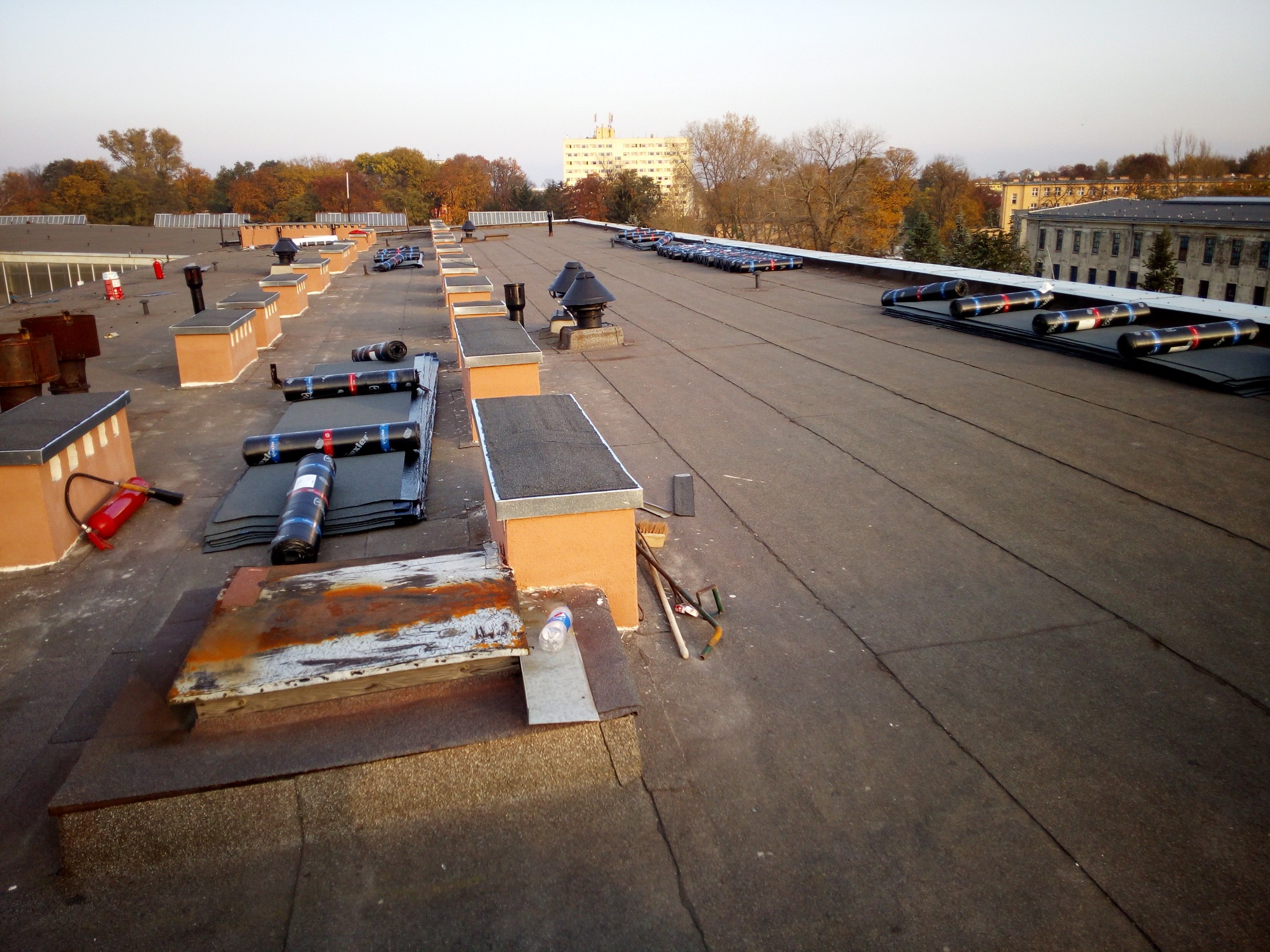 Rozległy, płaski dach pokryty papą termozgrzewalną w trakcie prac. Widoczne kominki wentylacyjne, zrolowane arkusze papy, narzędzia i budynki w tle.
