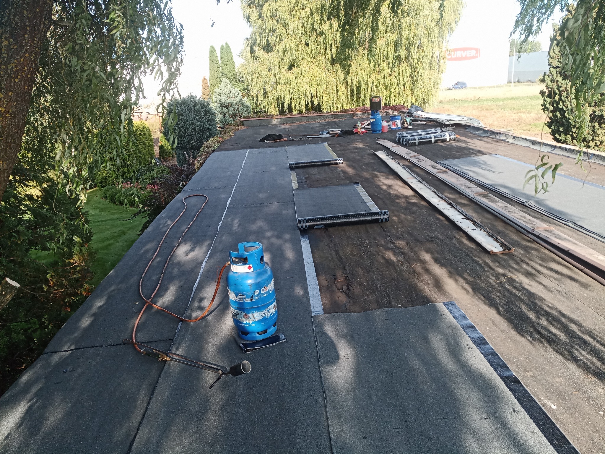Prace dekarskie na płaskim dachu: częściowo położona papa, butla gazowa z palnikiem, narzędzia dekarskie i rolki papy w tle.