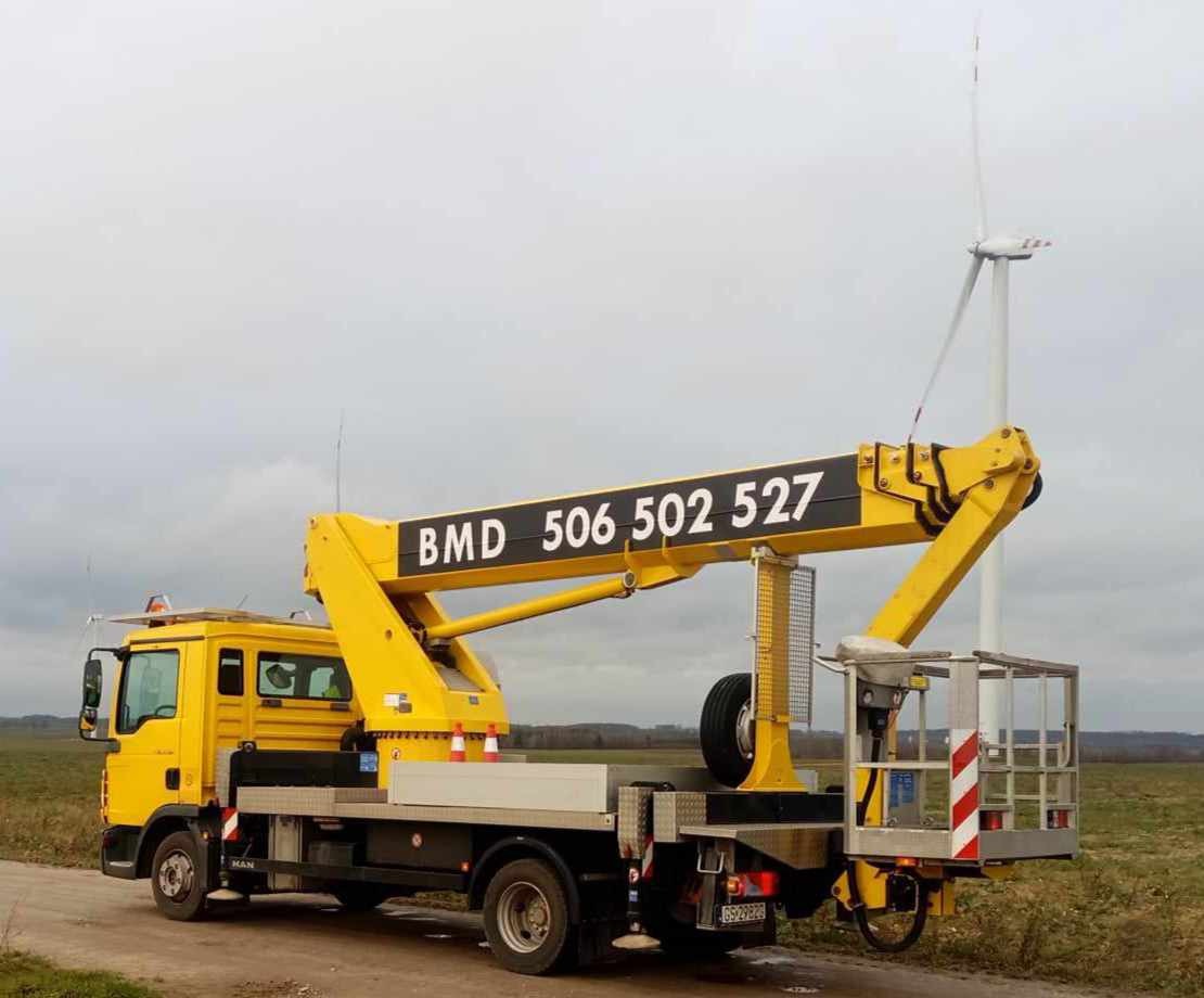 Żółty podnośnik koszowy MAN z wysięgnikiem z numerem telefonu BMD 506 502 527 na tle pochmurnego nieba i turbiny wiatrowej na polu.