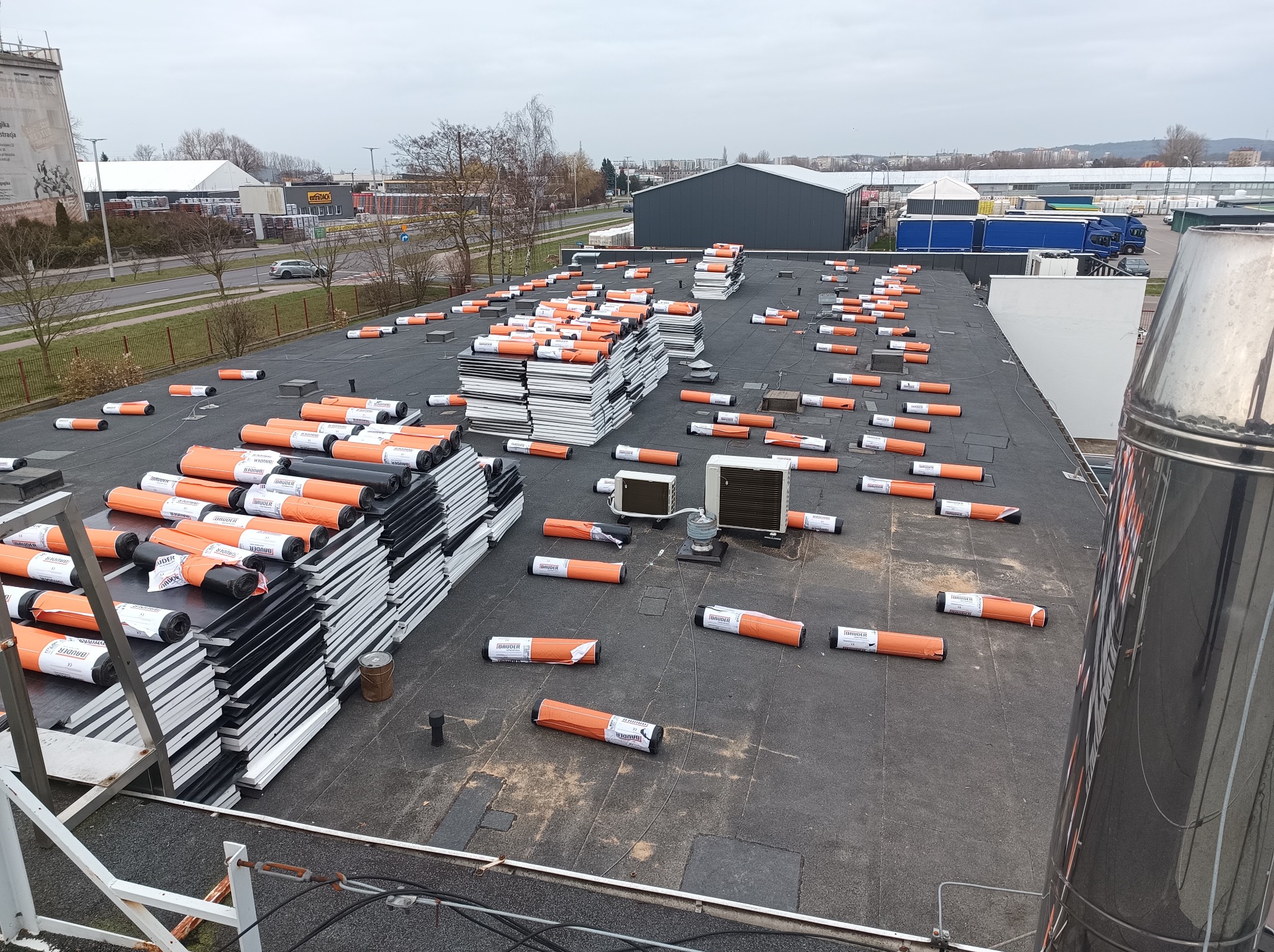 Widok z góry na płaski dach z rozłożonymi rolkami papy termozgrzewalnej marki Bauder w pomarańczowych opakowaniach oraz ułożonymi w stosy płytami izolacyjnymi z polistyrenu ekstrudowanego, w tle...