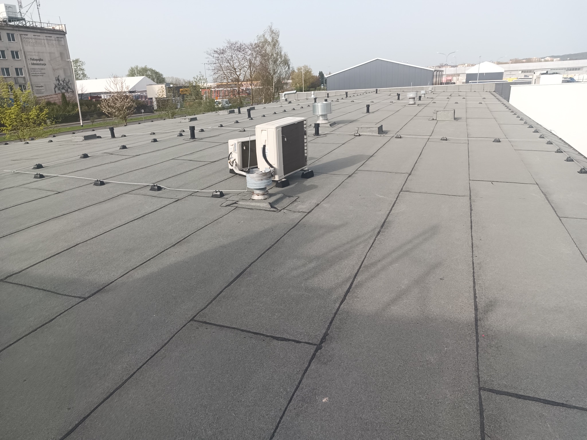 Płaski dach pokryty papą termozgrzewalną z widocznymi elementami wentylacyjnymi i klimatyzatorami, zamocowanymi na specjalnych podstawach, na tle budynków przemysłowych.