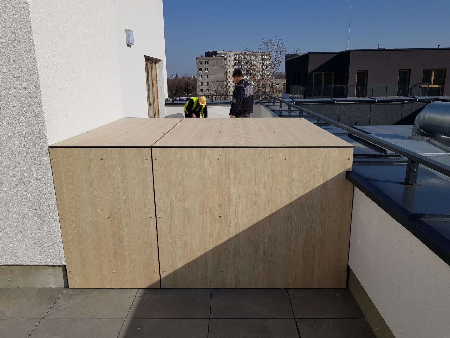 Zabudowa balkonu na dachu budynku wielorodzinnego, wykończona panelami imitującymi jasne drewno, z widocznymi dwoma pracownikami w tle, jeden w kamizelce odblaskowej.