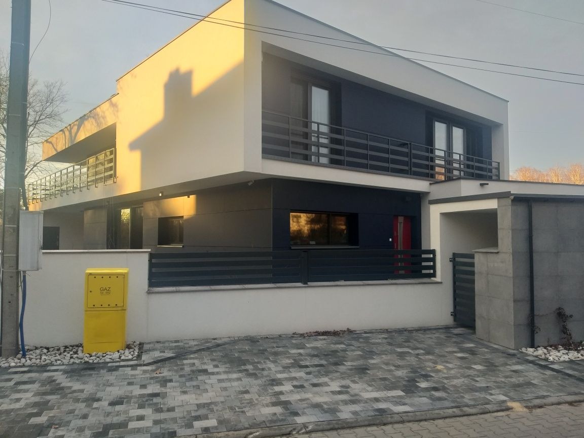 Nowoczesny, dwukondygnacyjny dom z płaskim dachem, wykończony w bieli i grafitowej szarości, z balkonem z metalową balustradą i żółtą skrzynką gazową na pierwszym planie.