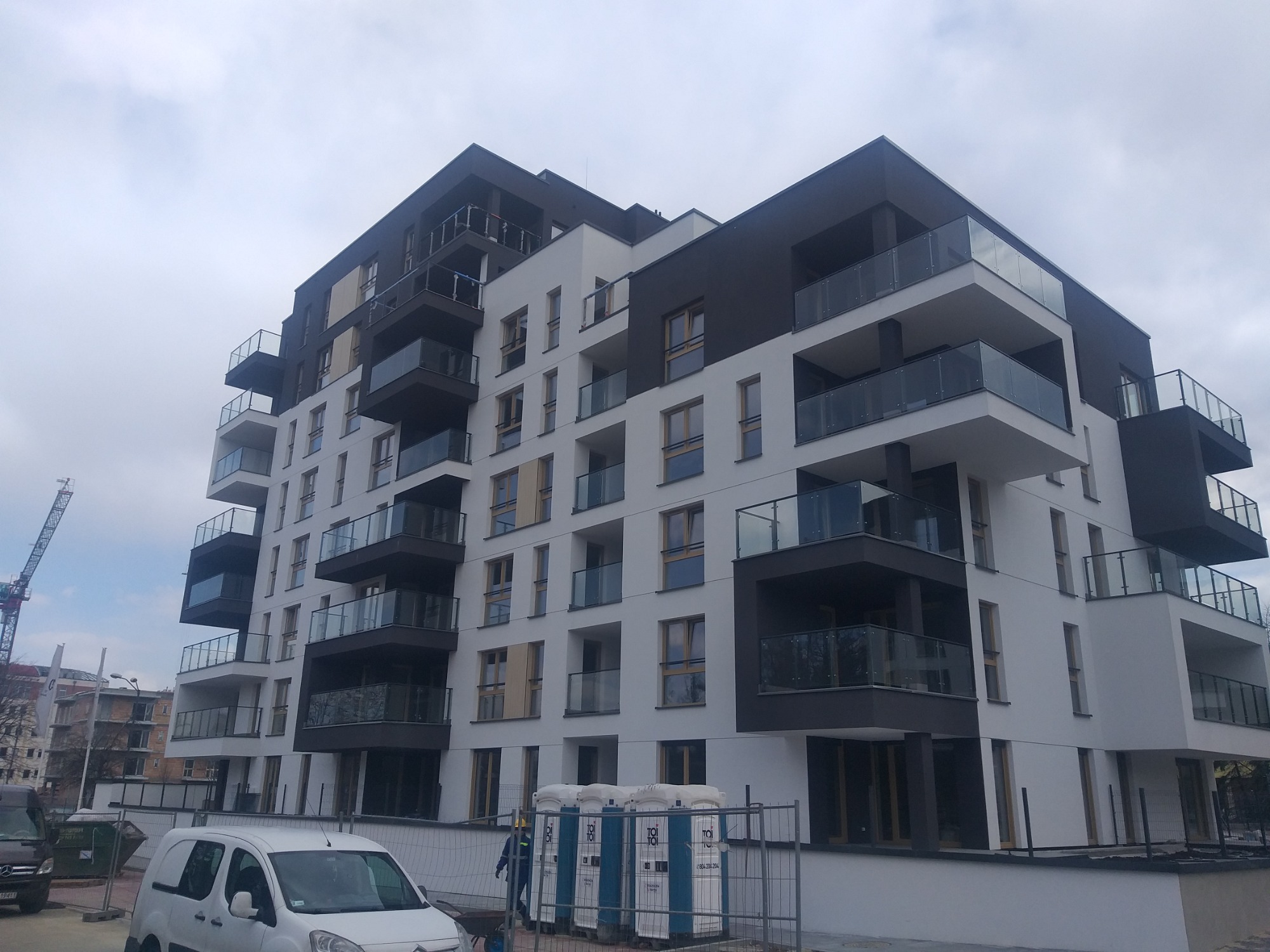 Nowoczesny budynek wielorodzinny z balkonami o szklanych balustradach, widoczne przenośne toalety na placu budowy i dźwig w tle.