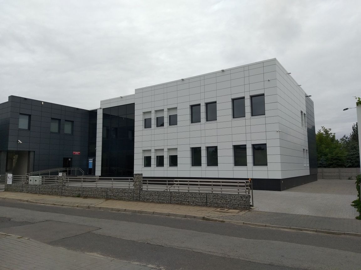 Nowoczesny budynek biurowy z elewacją z białych paneli i ciemnymi oknami, widoczny z poziomu ulicy w pochmurny dzień.