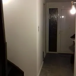 Korytarz z wykończonymi ścianami w kolorze białym, szare płytki podłogowe imitujące marmur, widoczne fragmenty schodów i drzwi wejściowych.