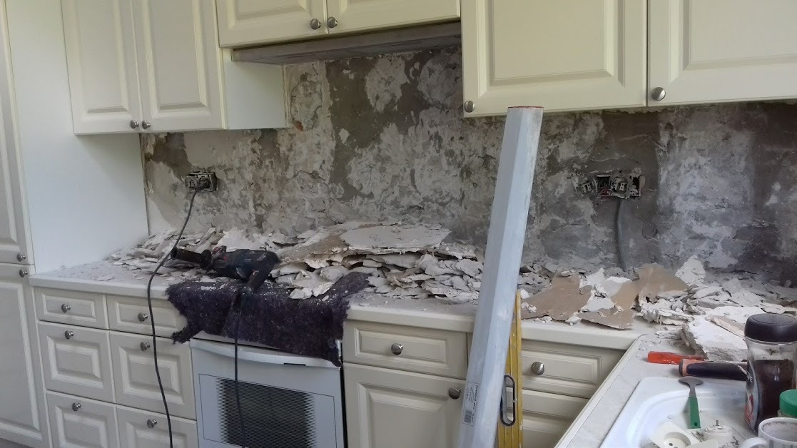 Demontaż starych płytek w kuchni, widoczne białe szafki i odsłonięta ściana z instalacją elektryczną, gruz na blacie i narzędzia.