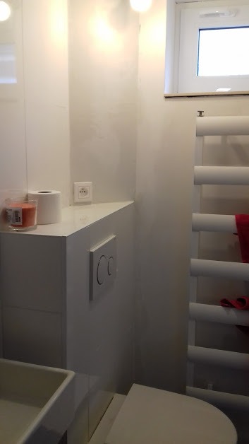 Biała łazienka z toaletą, grzejnikiem drabinkowym, półką z papierem toaletowym i świecą zapachową, oraz gładką ścianą z włącznikiem światła.