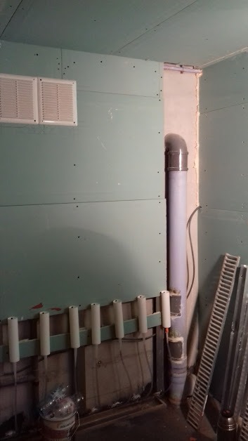 Ściana z płyt kartonowo-gipsowych przed szpachlowaniem, widoczne otwory po wkrętach, rura kanalizacyjna z kolankiem i grzejnik żeberkowy oparty o ścianę.