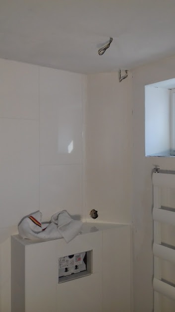 Wnętrze łazienki w trakcie prac: białe płytki na ścianach, wystające kable z sufitu, grzejnik drabinkowy i zabudowa podtynkowego WC z widoczną instalacją elektryczną, zakryta roboczą szmatką.