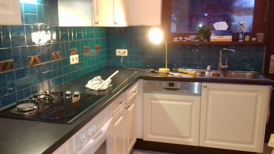 Kuchnia z turkusowymi płytkami na ścianie, czarnym blatem, białymi szafkami i wbudowaną płytą grzewczą. Widoczne narzędzia i akcesoria sugerują etap prac wykończeniowych.