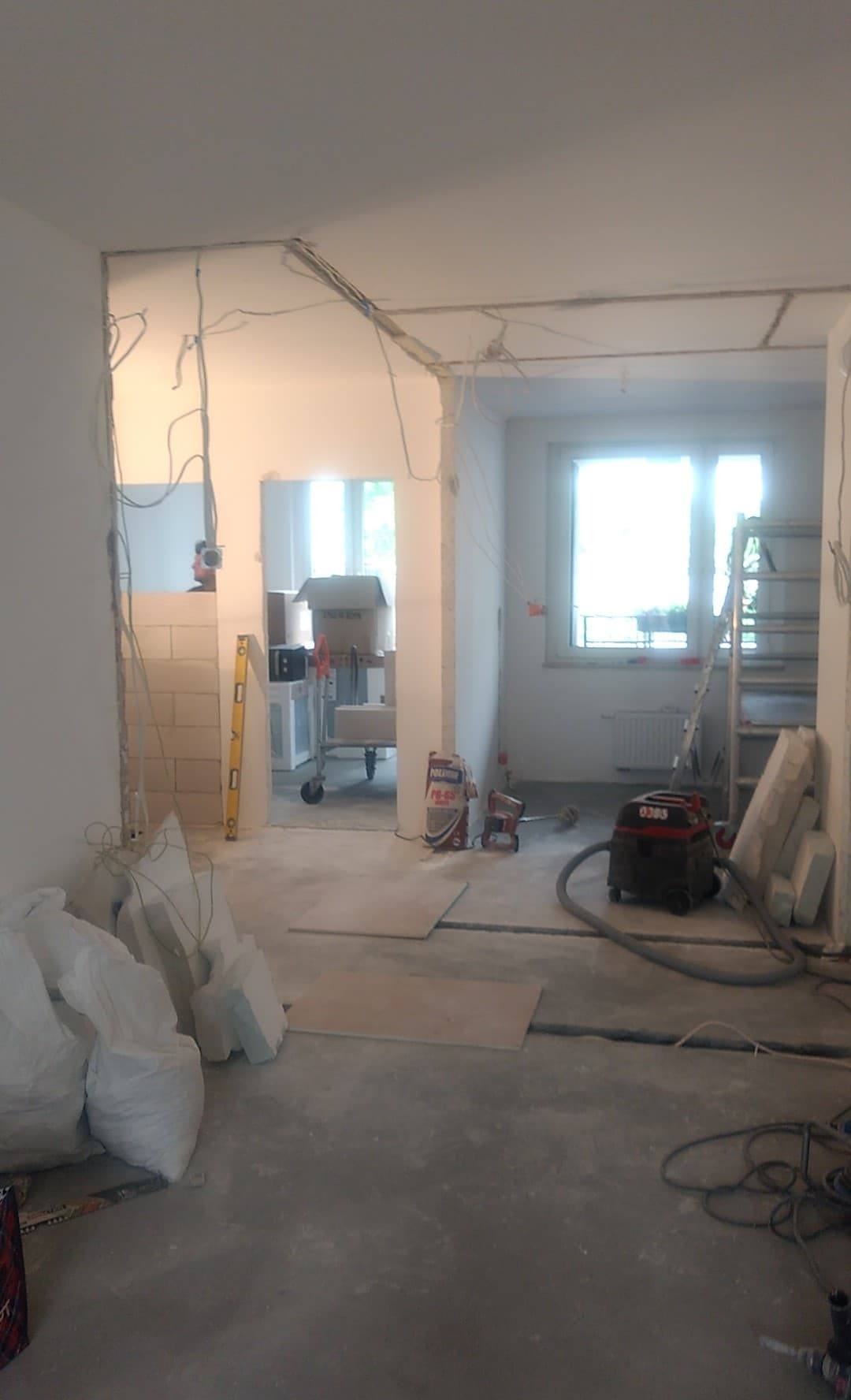 Wnętrze mieszkania w stanie deweloperskim w trakcie wyburzania ścian i zmiany instalacji, widoczne worki z materiałem, poziomica, odkurzacz przemysłowy i drabina.