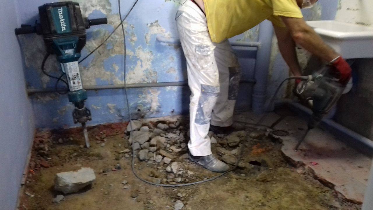 Demontaż starej posadzki w łazience za pomocą młota udarowego Makita, widoczne gruz i fragmenty starej instalacji sanitarnej.