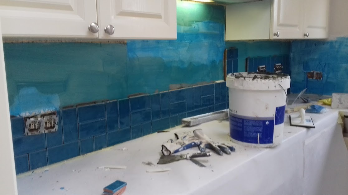 Montaż niebieskich płytek w kuchni w trakcie realizacji, widoczny kubeł z zaprawą, narzędzia glazurnicze i fragmenty niezabezpieczonych ścian.