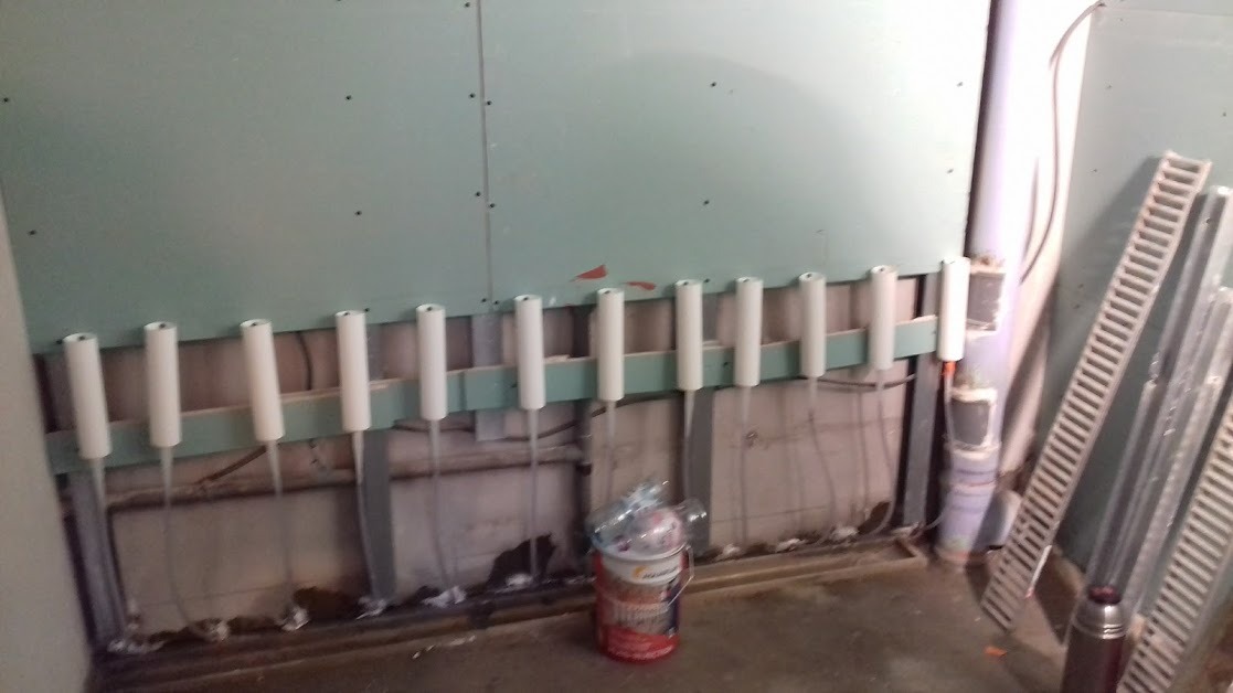 Niedokończona ściana z płyt gipsowo-kartonowych z widocznymi rurami grzewczymi zabezpieczonymi białymi osłonami, obok stoi wiadro z farbą i aluminiowe profile.