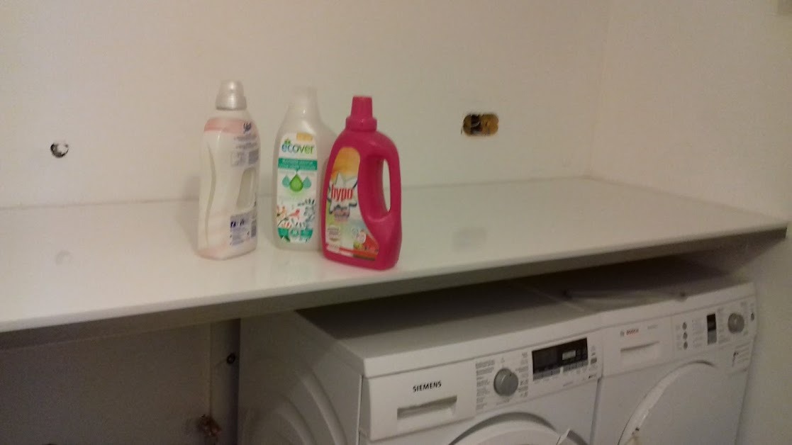 Biały blat nad pralkami Siemens i Bosch, z trzema butelkami detergentów: różowy, zielony Ecover i biały, widoczne gniazdko elektryczne bez osłony.