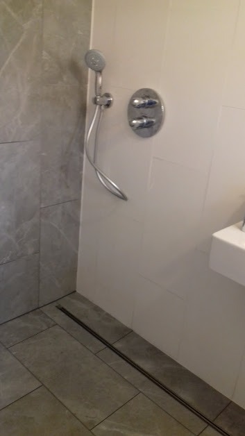 Nowoczesna łazienka z szarymi płytkami imitującymi marmur, odpływem liniowym i chromowaną armaturą prysznicową na białej ścianie.