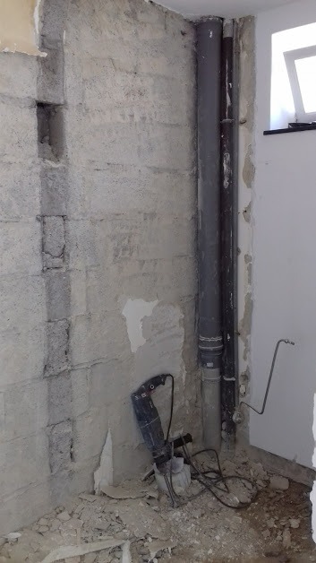 Surowa ściana z betonu komórkowego w trakcie remontu łazienki, widoczne rury kanalizacyjne i młot udarowy leżący na podłodze.