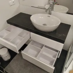 Nowoczesna łazienka z blatem imitującym czarny marmur, owalną białą umywalką nablatową i szafką z szufladami z przegródkami na akcesoria.