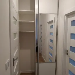 Biała szafa wnękowa z lustrem w wąskim przedpokoju, widoczne półki wewnętrzne i odbicie kolejnych drzwi w lustrze.