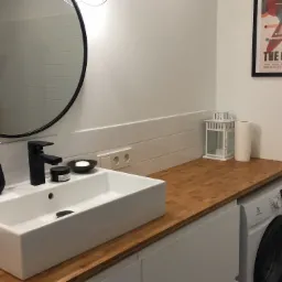 Nowoczesna łazienka z drewnianym blatem, białą, prostokątną umywalką nablatową, czarną armaturą, okrągłym lustrem i pralką pod blatem. Na blacie dekoracje: lampion i pojemnik na papier toaletowy.