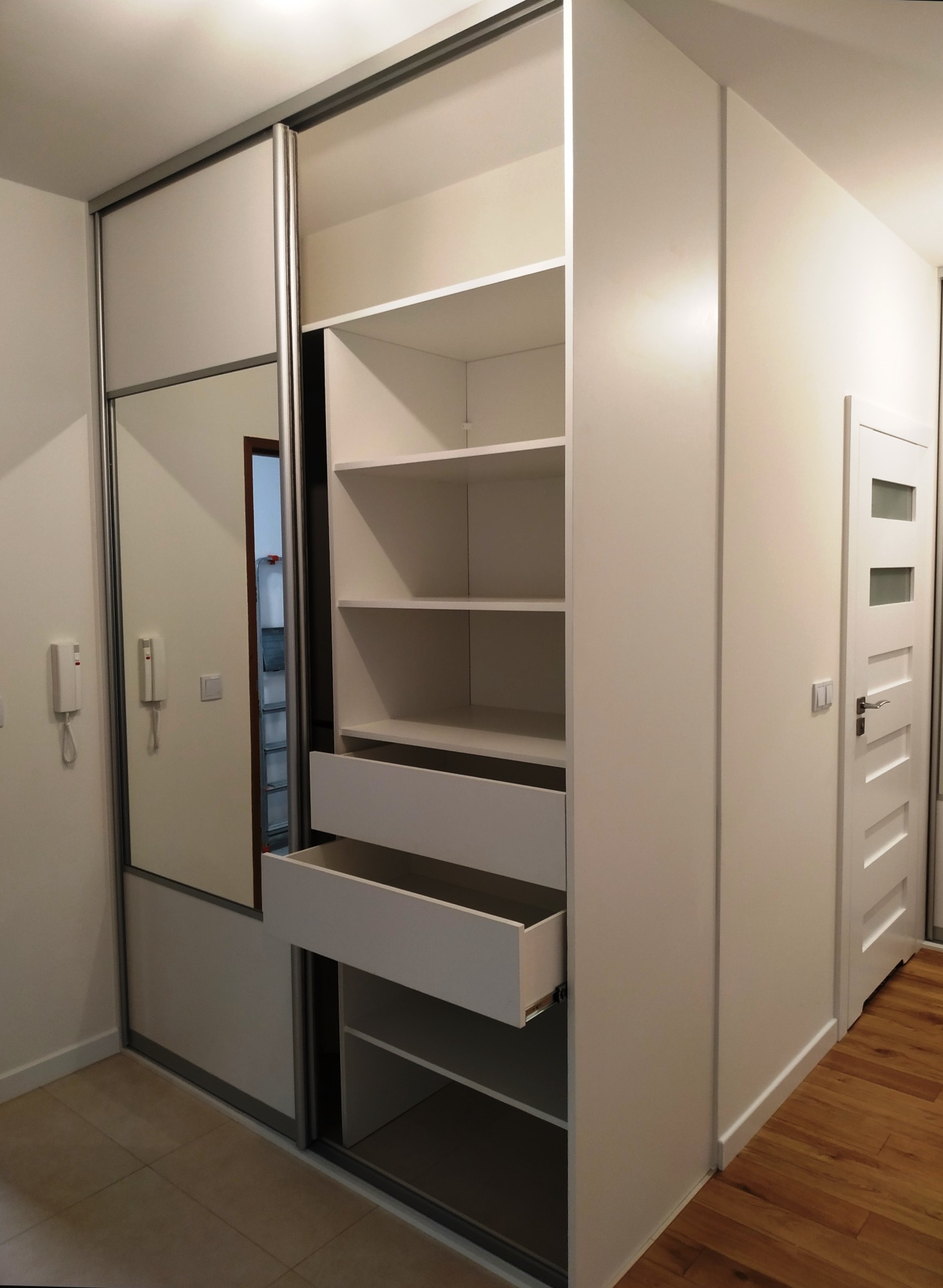 Biała szafa wnękowa z przesuwnymi drzwiami z lustrem, częściowo otwarta, ukazująca półki i wysuniętą szufladę, widoczna drewniana podłoga i białe drzwi wejściowe.