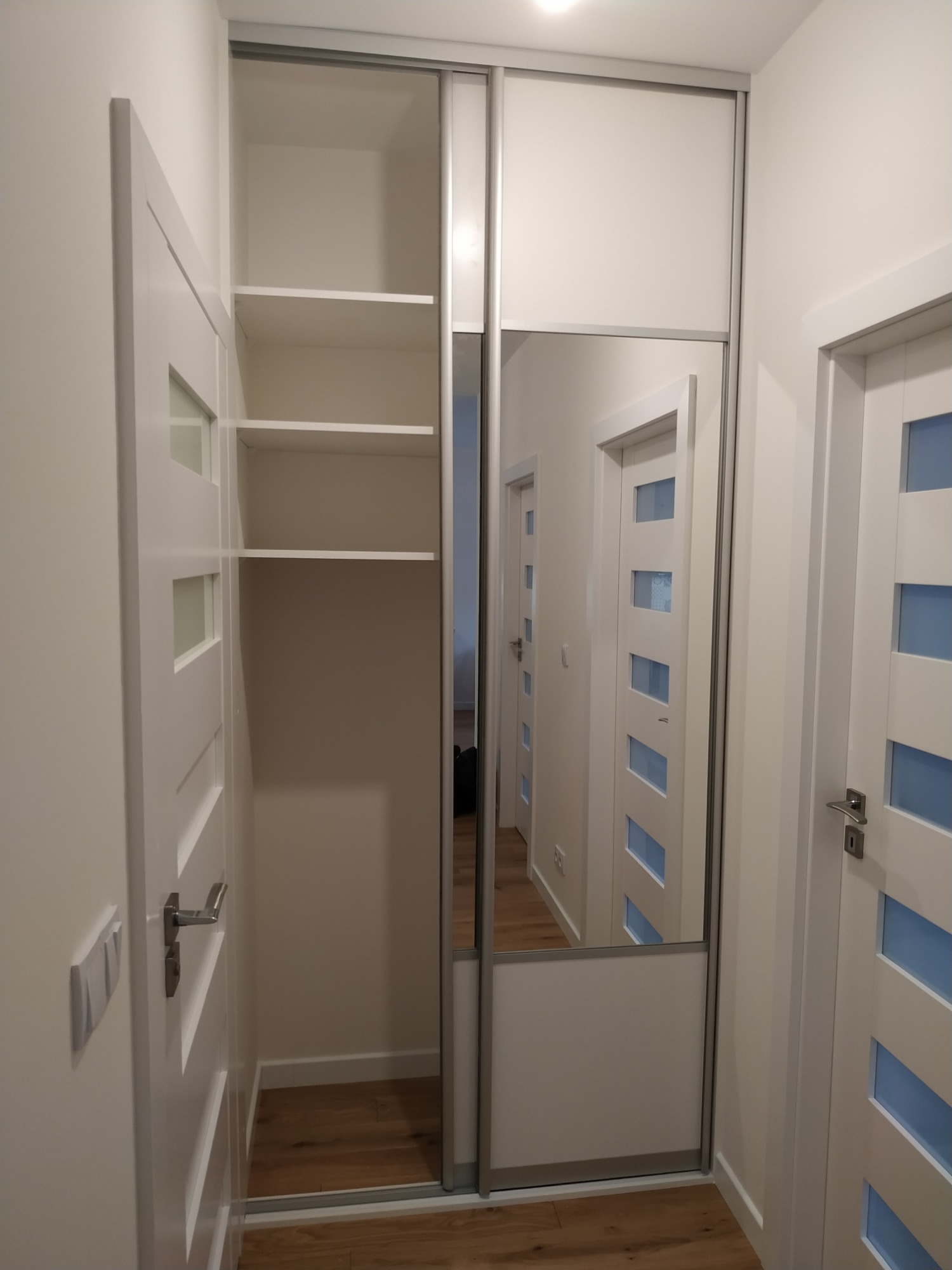 Biała szafa wnękowa z lustrem w wąskim przedpokoju, widoczne półki wewnętrzne i odbicie kolejnych drzwi w lustrze.