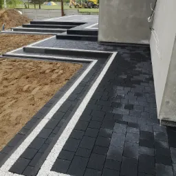 Schody i chodnik z ciemnej kostki brukowej z jasnymi obrzeżami, w tle plac budowy z koparką.